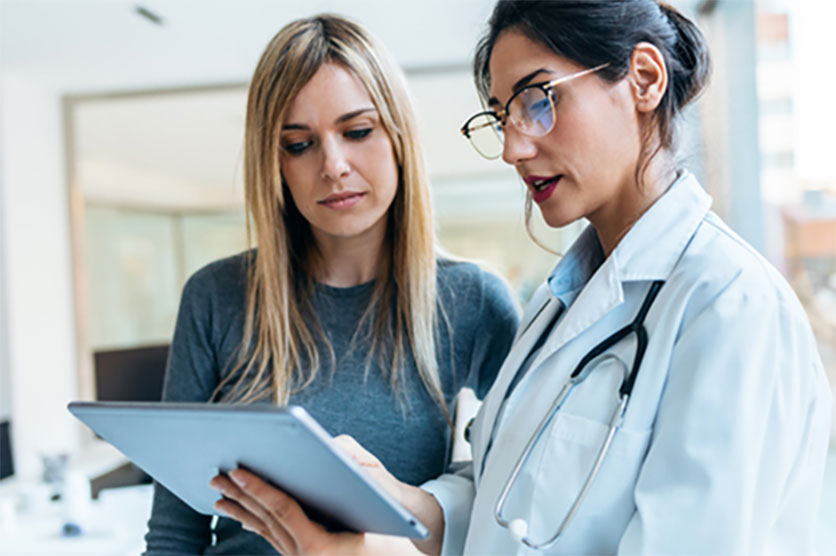 Two women looking at a tablet. The one on the right of the image is weird a white lab coat with a stethoscope hanging over her shoulder.